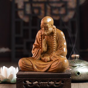 May include: A wooden statue of a seated figure with a long beard and a beaded necklace. The statue is carved in a detailed style and has a warm brown color. The statue is sitting on a wooden stand with intricate carvings.