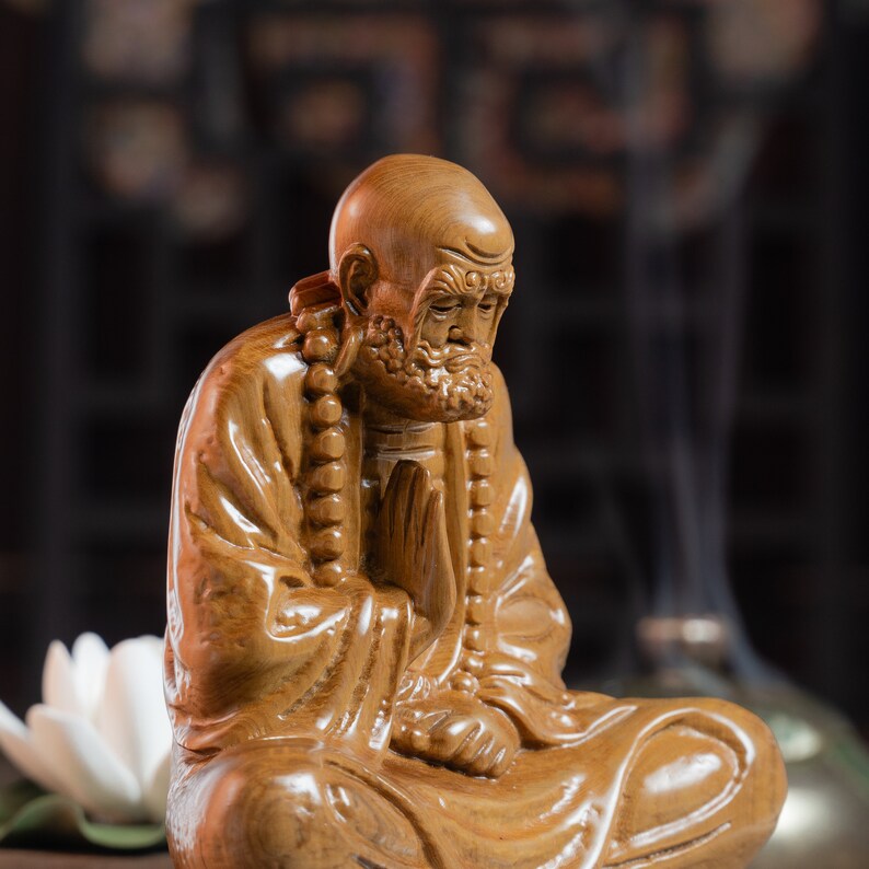 May include: A wooden sculpture of a seated figure with a long beard and a serene expression. The figure is wearing a robe and has a beaded necklace around its neck.