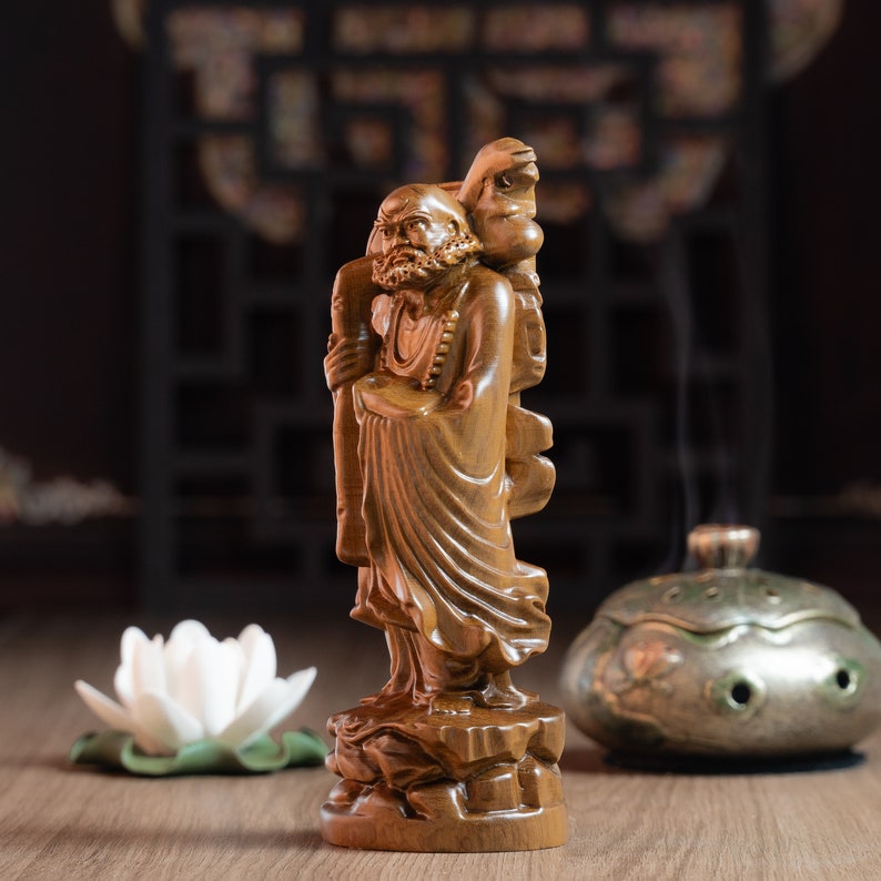 May include: A wooden figurine of a smiling, bearded man holding a staff. The figurine is carved in a detailed style and has a natural wood finish. The man is wearing a long robe and has a peaceful expression.