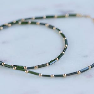 May include: A delicate necklace featuring dark green, rectangular beads interspersed with small, gold-colored beads. The necklace is displayed on a white marble surface, showcasing its elegant design and color contrast.