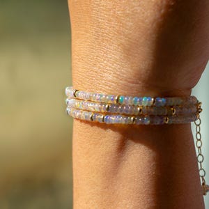 Puede incluir: Un brazalete de capas con pequeñas cuentas de ópalo iridiscente y detalles dorados. El brazalete tiene una delicada extensión de cadena.