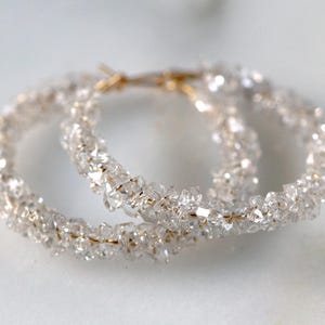 May include: A pair of gold hoop earrings adorned with clear, sparkling crystals. The crystals have an irregular shape, providing a textured look. The earrings are presented on a white surface, accentuating their sparkle.