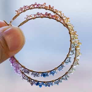 May include: A pair of gold hoop earrings with a rainbow of small gemstones. The gemstones are arranged in a gradient from pink to yellow to blue.