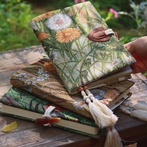May include: A stack of three floral-patterned journals with a nature-inspired design. The covers feature dandelions, daisies, and other wildflowers in shades of green, yellow, and white. Each journal has a unique closure and tassel.