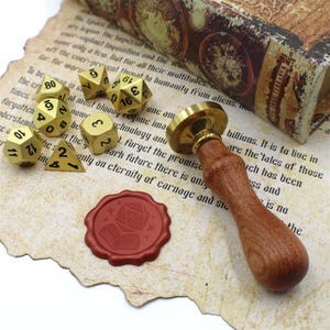 May include: A collection of gold-colored polyhedral dice, a wooden-handled wax stamp, and a red wax seal with a D20 design, all arranged on a vintage paper background. A book is visible in the background.