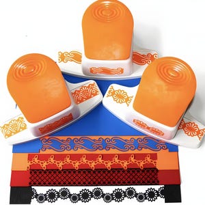 May include: Three orange and white border punch tools with different designs. The tools are on a blue surface with samples of the borders punched in orange, red, and black paper.