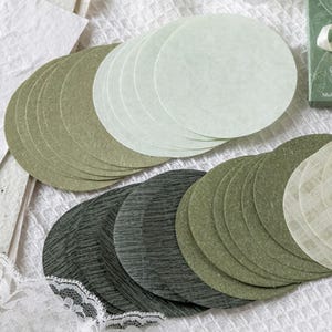May include: A selection of circular paper coasters in shades of green and white. The coasters have a textured finish, with some displaying a delicate pattern. They are arranged in stacks, suggesting they are ready for use.