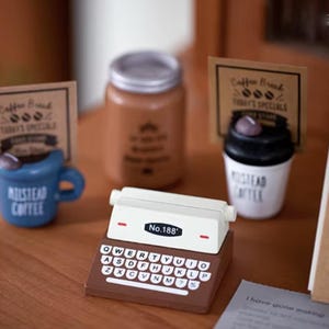 Puede incluir: Una máquina de escribir en miniatura con la parte superior blanca y la base marrón, con un teclado con letras y números. El texto "No.188" es visible. Otros artículos incluyen una taza azul, una taza de café y un tarro.