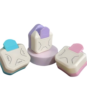 Puede incluir: Tres perforadoras de papel redondeadoras de esquinas en blanco, con detalles en azul, morado y rosa. Cada perforadora tiene un radio de esquina diferente, etiquetado R 4mm, R 7mm y R 10mm. Las perforadoras están diseñadas para manualidades y álbumes de recortes.