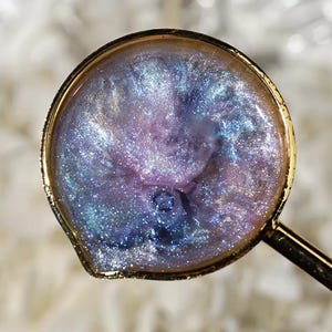 Puede incluir: Un objeto redondo de resina dorada con un mango, lleno de una mezcla arremolinada de purpurina morada, azul y plateada. La purpurina crea un efecto de galaxia, con una superficie brillante y reflectante.