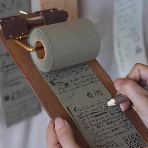 Puede incluir: Un bloc de notas de madera con un rollo de papel verde y un clip de metal dorado. El bloc de notas tiene notas manuscritas en el papel. Una persona está escribiendo en el papel con un lápiz.