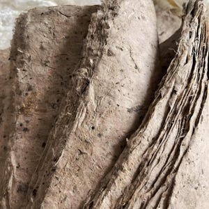 Peut inclure: Gros plan sur des feuilles de papier faites à la main. Le papier a une surface rugueuse et texturée avec des fibres et des mouchetures visibles. La couleur est un beige naturel et terreux. Les bords des feuilles sont inégaux, suggérant un processus artisanal.