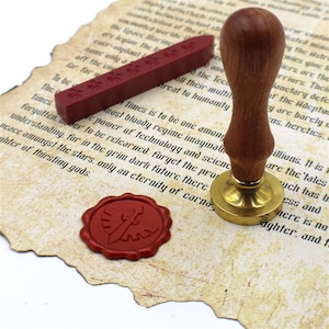 Puede incluir: Un juego de sellado de cera de época con un sello con mango de madera y base de latón, una barra de cera roja y un sello de cera estampado. El juego está sobre un papel tipo pergamino con texto.