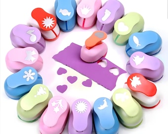 May include: A set of 14 colorful plastic paper punchers with different shapes, including a star, a heart, a snowflake, a crown, a fish, a leaf, a sun, a tree, a dolphin, a bow, a musical note, a candy, a footprint, and a maple leaf. The punchers are arranged in a circle around a purple sheet of paper with cut-out hearts.