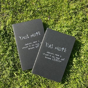Peut inclure: Deux cahiers noirs avec du texte manuscrit blanc, posés sur de l'herbe verte. L'un dit "YAOI NOTE" et l'autre "YURI NOTE". Le texte est une référence humoristique à un anime populaire.