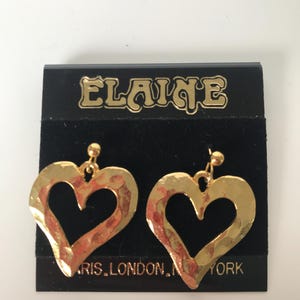 May include: A pair of gold-colored heart-shaped earrings with a hammered texture. The earrings hang from small gold studs. The earrings are displayed on a black card with the name "Elaine" in gold lettering.