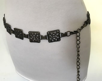 May include: A brown metal belt with a chain and decorative square links. The belt is worn around the waist of a person.