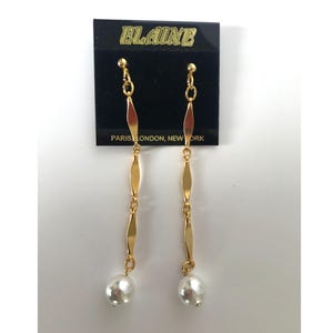 May include: A pair of gold dangle earrings with a pearl at the bottom. The earrings are on a black card with the text "ELAINE PARIS LONDON, NEW YORK".