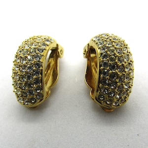 May include: A pair of gold tone clip-on earrings with clear rhinestones. The earrings are shaped like a half-circle and are covered in rhinestones.
