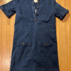 May include: A dark blue denim dress with short sleeves, a button-up front, and two patch pockets. The dress has a small label that says "DENIM" on the front.