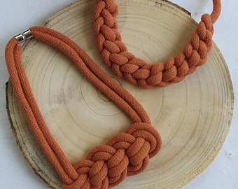 Puede incluir: Dos collares de cuerda marrón con un diseño anudado. Los collares se muestran sobre una superficie de madera.