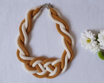 Puede incluir: Un collar trenzado hecho de dos hebras de cuerda, una de color amarillo mostaza y otra blanca. El collar está anudado en el centro y tiene cierres plateados.