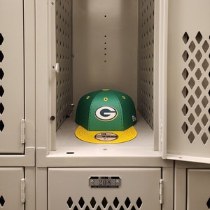 May include: A green and yellow baseball cap with a green "G" logo and the text "New Era" on the side. The cap is in a gray locker with a metal tag that reads "LYON 208".
