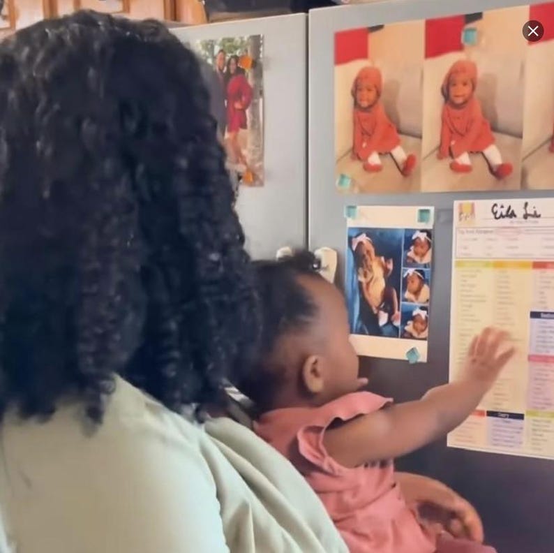 Puede incluir: Una mujer con el pelo largo y negro rizado sostiene a un ni&ntilde;o peque&ntilde;o en sus brazos. El ni&ntilde;o est&aacute; mirando una nevera cubierta de imanes y fotos. Una foto muestra a una ni&ntilde;a peque&ntilde;a con un vestido rojo y un sombrero blanco. La nevera tambi&eacute;n tiene un calendario con la palabra "Eid" escrita en &eacute;l.