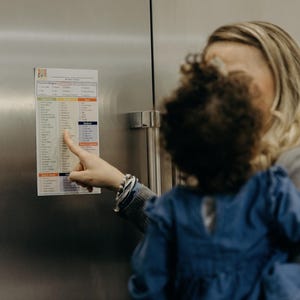 Puede incluir: Una mujer se&ntilde;ala un im&aacute;n de nevera con una lista que dice "The Food Allowance". La lista est&aacute; dividida en categor&iacute;as, que incluyen frutas, verduras, cereales y prote&iacute;nas. La mujer lleva a un ni&ntilde;o peque&ntilde;o en brazos.