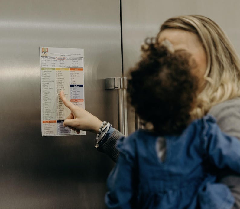 Puede incluir: Una mujer se&ntilde;ala un im&aacute;n de nevera con una lista que dice "The Food Allowance". La lista est&aacute; dividida en categor&iacute;as, que incluyen frutas, verduras, cereales y prote&iacute;nas. La mujer lleva a un ni&ntilde;o peque&ntilde;o en brazos.