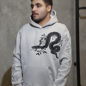 May include: A light grey hooded sweatshirt with a black and white embroidered snake design on the front. The snake is wrapped around a branch with flowers and leaves. The sweatshirt has a kangaroo pocket and a drawstring hood.