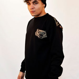 Puede incluir: Una sudadera negra con una gran cara de animal bordada en la parte delantera. La sudadera tiene un dise&ntilde;o floral en el hombro derecho. La persona que lleva la sudadera lleva una boina negra en la cabeza y un tatuaje en la mu&ntilde;eca.
