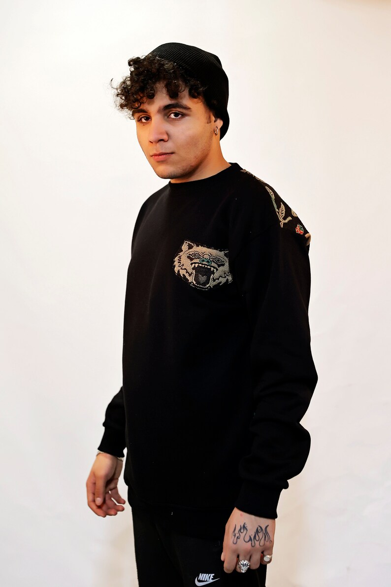 Puede incluir: Una sudadera negra con una gran cara de animal bordada en la parte delantera. La sudadera tiene un dise&ntilde;o floral en el hombro derecho. La persona que lleva la sudadera lleva una boina negra en la cabeza y un tatuaje en la mu&ntilde;eca.