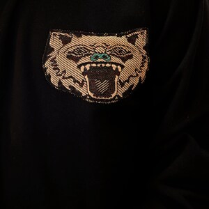 Puede incluir: Sudadera negra con una cara de animal bordada en marr&oacute;n y blanco con ojos verdes. El animal est&aacute; gru&ntilde;endo con la boca abierta.