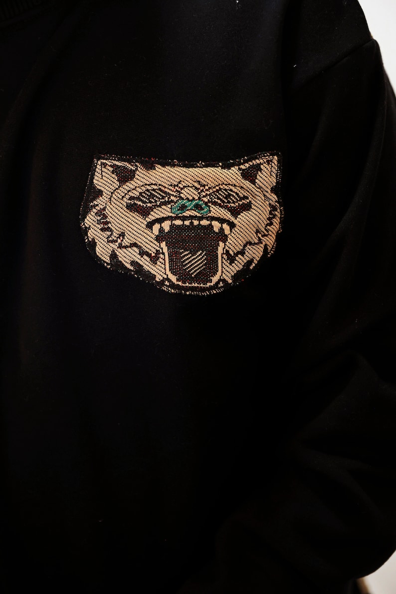 Puede incluir: Sudadera negra con una cara de animal bordada en marr&oacute;n y blanco con ojos verdes. El animal est&aacute; gru&ntilde;endo con la boca abierta.