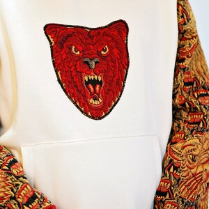 May include: White hoodie with a red embroidered bear face on the front and red and gold embroidered animal faces on the sleeves.