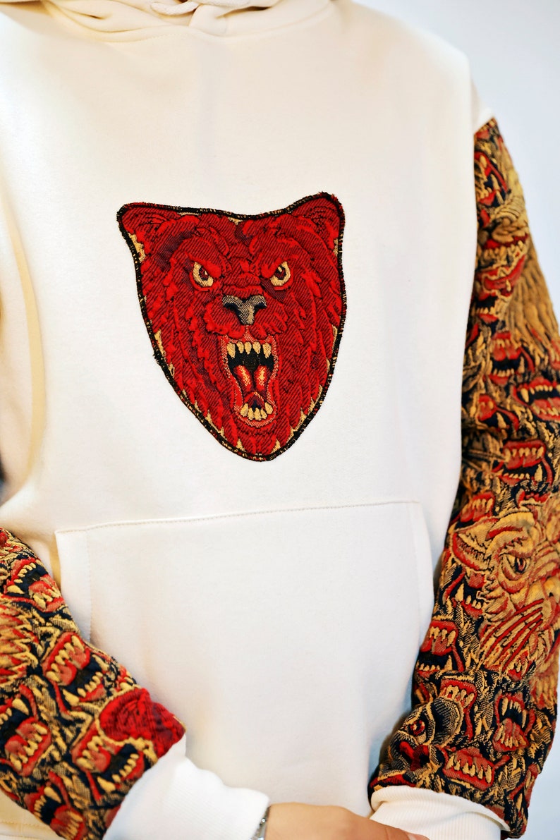 May include: White hoodie with a red embroidered bear face on the front and red and gold embroidered animal faces on the sleeves.