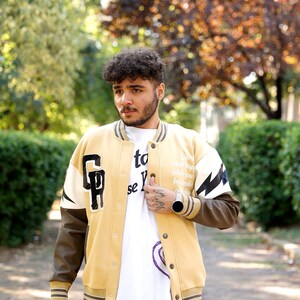 May include: A man wearing a tan and brown varsity jacket with white and black lightning bolt and letter patches. The jacket has a zipper closure and a white collar. The man is wearing a white t-shirt with black text and a black watch.