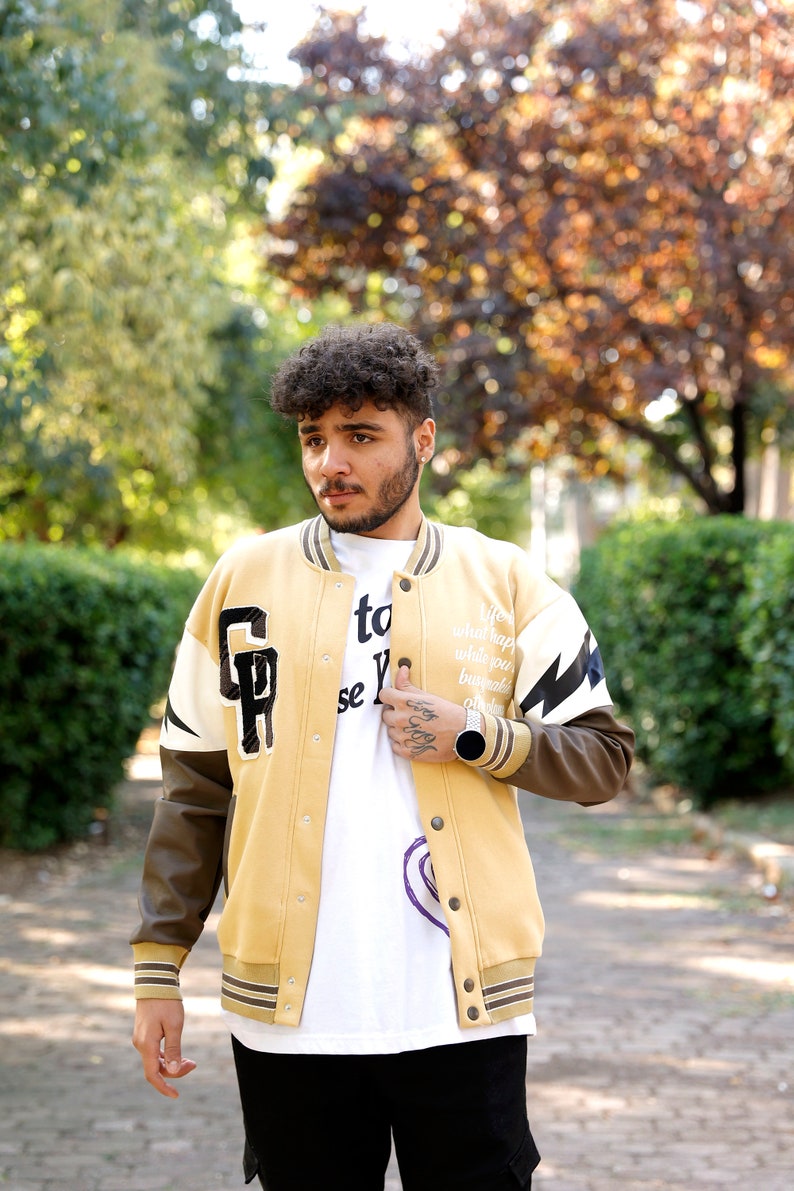 May include: A man wearing a tan and brown varsity jacket with white and black lightning bolt and letter patches. The jacket has a zipper closure and a white collar. The man is wearing a white t-shirt with black text and a black watch.