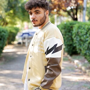 May include: A man wearing a beige varsity jacket with brown leather sleeves and a white lightning bolt graphic. The jacket has a white zipper and white text on the front.