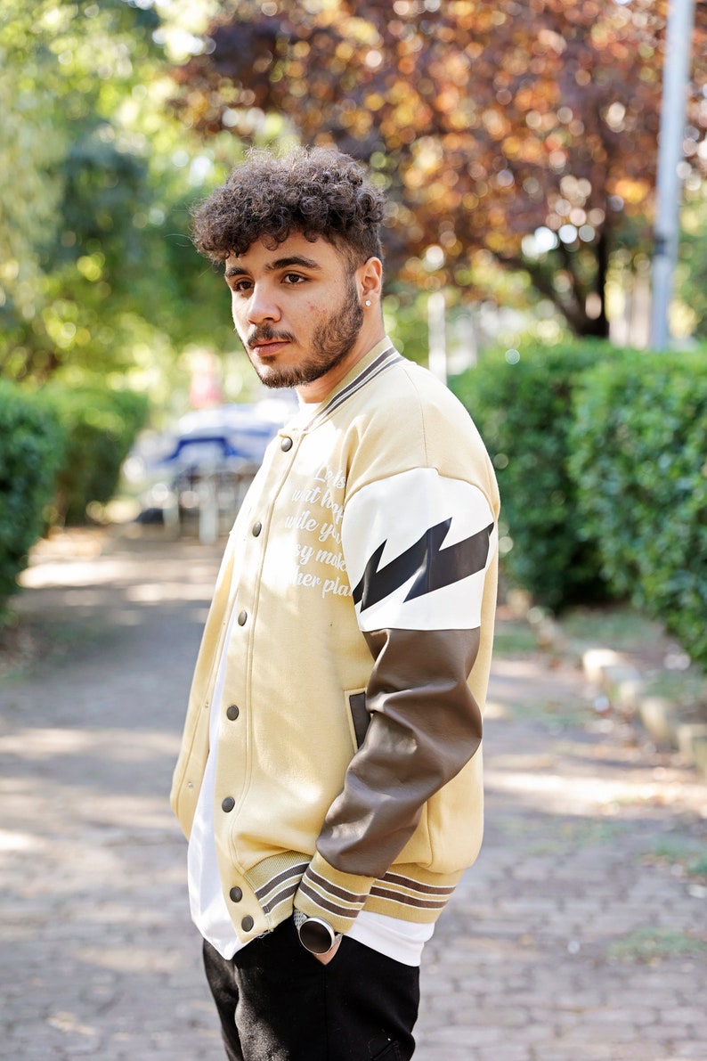 May include: A man wearing a beige varsity jacket with brown leather sleeves and a white lightning bolt graphic. The jacket has a white zipper and white text on the front.