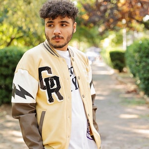 May include: A person wearing a beige varsity jacket with brown leather sleeves. The jacket has a large "GR" patch on the left side and a white lightning bolt design on the right sleeve. The person is wearing a white t-shirt underneath the jacket.