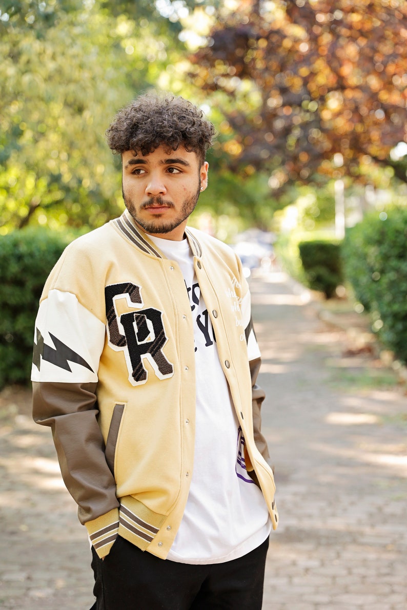 May include: A person wearing a beige varsity jacket with brown leather sleeves. The jacket has a large "GR" patch on the left side and a white lightning bolt design on the right sleeve. The person is wearing a white t-shirt underneath the jacket.