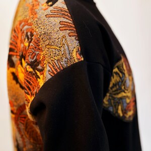 May include: Black sweatshirt with a colourful embroidered design on the left shoulder and sleeve. The design features a mix of orange, yellow, and blue colours.