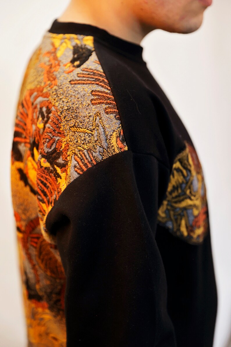 May include: Black sweatshirt with a colourful embroidered design on the left shoulder and sleeve. The design features a mix of orange, yellow, and blue colours.