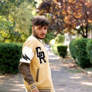 May include: A young person wearing a yellow and brown varsity jacket with a black and white lightning bolt design on the sleeve and the letters "GR" on the chest. The jacket has a white ribbed collar and cuffs.
