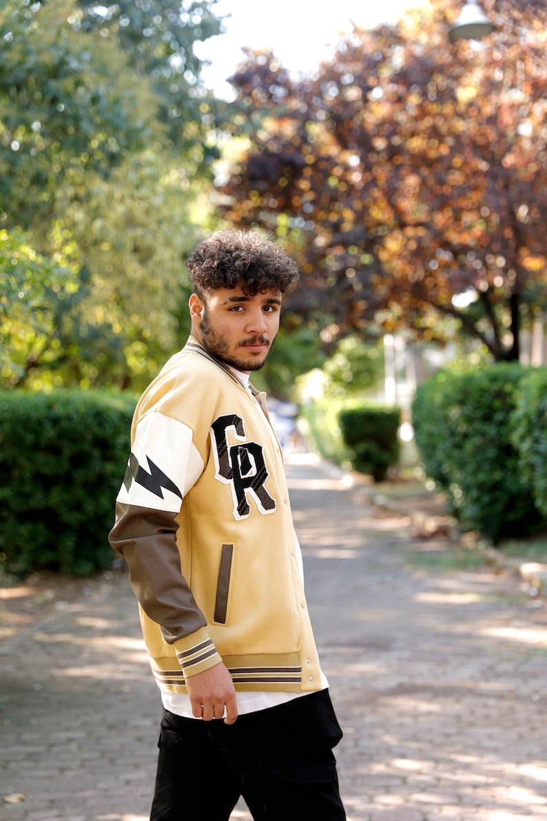 May include: A young person wearing a yellow and brown varsity jacket with a black and white lightning bolt design on the sleeve and the letters "GR" on the chest. The jacket has a white ribbed collar and cuffs.