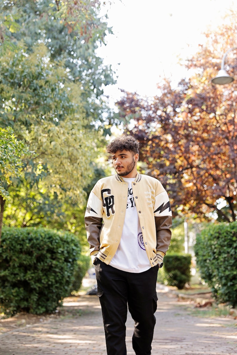 May include: A person wearing a beige and brown varsity jacket with white and black accents. The jacket has the letters "GR" on the left side and the text "NYC" on the right side. The person is wearing a white t-shirt with a graphic design and black pants.