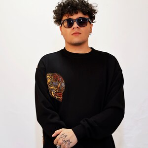 May include: A black sweatshirt with a colourful embroidered skull on the left sleeve. The model is wearing black sunglasses and has curly hair.