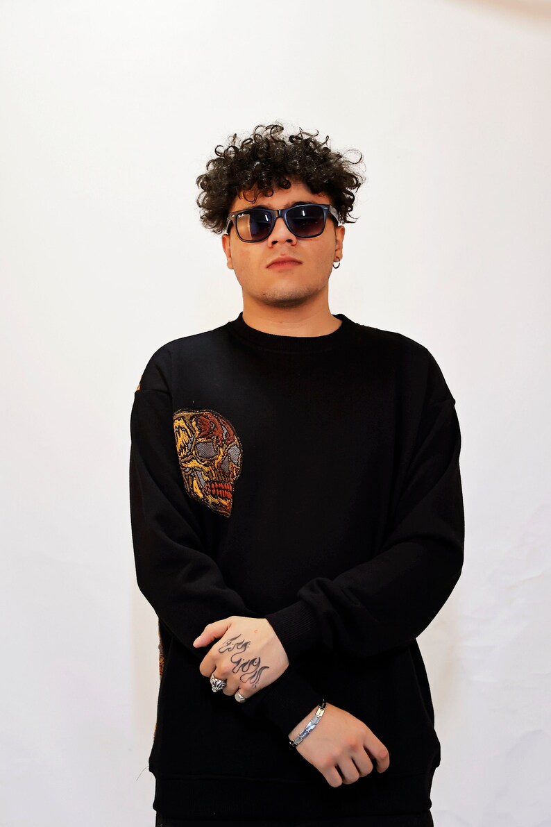 May include: A black sweatshirt with a colourful embroidered skull on the left sleeve. The model is wearing black sunglasses and has curly hair.